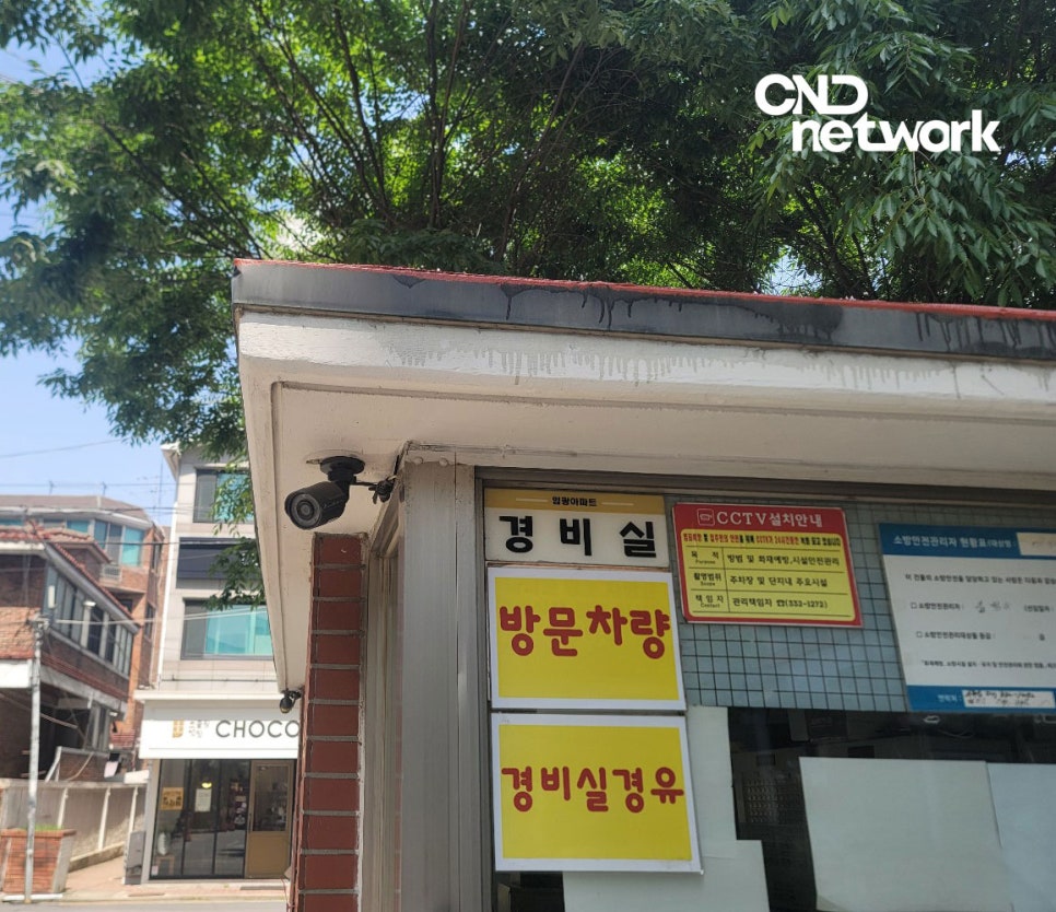 아파트 경비실 처마 밑에 부착된 실외형 카메라, 방문차량 및 경비실 안내판과 함께 촬영된 현장